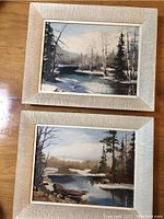 Photo showing two small original oil paintings in light wooden frames on a wooden surface. One painting depicts a winter landscape with snow and a river, the other an autumn river scene with trees.