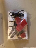 Red electric soldering tool with coiled black power cord, metal tips, and small container, inside plastic packaging placed on concrete floor.