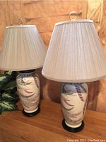 Set of two ceramic-based table lamps with leaf design and pleated fabric shades with ring finials, placed against a beige background with leaf plant on side.