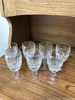 Seven Waterford sherry glasses with detailed cut crystal design, short stems arranged in rows on a wooden shelf.