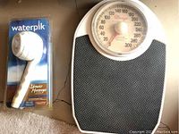 Mechanical dial bathroom scale with orange dial next to Waterpik shower massager in original packaging, both placed on carpeted floor.
