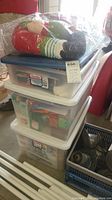 Stacked plastic storage tubs containing various Christmas decorations with a stuffed elf decoration on top in plastic bag