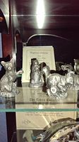 Photo showing several pewter woodland animal figurines on glass shelves, including bear, raccoon, and squirrel with a certificate visible behind them.