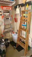 Two ladders hanging in a garage corner: one aluminum Little Giant Boost ladder with orange accents and one 6ft wooden step ladder with natural wood finish.