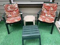 Two green plastic patio chairs with floral cushions, one beige plastic small table, and one green plastic slatted table arranged on green outdoor carpet.