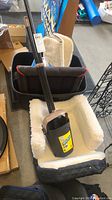 Poop scooper with long handle resting on a small pet bed with white fleece lining and black quilted exterior, along with black storage bin and cardboard boxes near items.