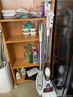 Wide view showing two Shark steam mops placed side by side next to a wooden shelf holding multiple cleaning supplies on top three shelves.