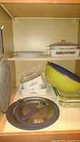 Shelf with stacked Fire King Corning Cornflower white glass casserole dishes, yellow mixing bowl, clear Pyrex dish, and metal grill basket with grease screens underneath