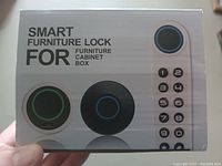 Image showing front view of the box packaging with text 'SMART FURNITURE LOCK FOR FURNITURE CABINET BOX' and images of the lock featuring different colored illuminated rings and keypad numbers.