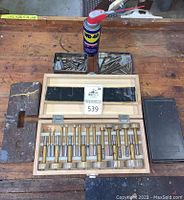 Wooden case open showing brass Forstner drill bits arranged inside with a can of WD-40 and two metal trays of bits behind.