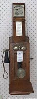 Frontal view of oak wall phone mounted on wallpapered wall showing dual brass bells, handset, mouthpiece, and attached Baker Brothers Groceries notepad