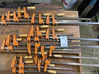 Overview of 16 orange Jorgensen steel bar clamps with wooden handles on a wooden workbench