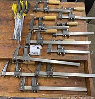Top-down view showing ten assorted metal clamps arranged on a wooden surface along with a wrapped set of needle nose pliers with yellow and black handles.