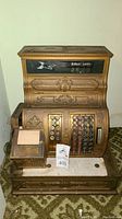Full front view showing the complete cash register with display, keys, and base on patterned carpet.