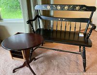 Black wooden bench with floral painted designs on upper backrest and middle back rail, featuring spindled back and armrests, visible carpet floor and window.