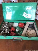 Open metal box revealing Stanlo Toy Erector set components including metal strips, nuts, bolts, and various toy parts.
