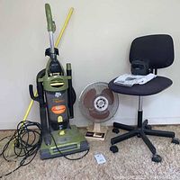 Full lot showing Dirt Devil vacuum, a yellow broom leaning behind it, Cool Breeze fan, and black rolling office chair against a plain white wall.