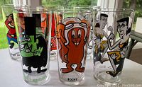 Photo showing six of the eight vintage cartoon character pint glasses, featuring Dudley Do-Right, Rocky, Woody Woodpecker, and others arranged on white surface near window.