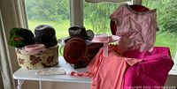 Wide view showing multiple vintage women's hats in various colors and styles, the Resistol cowboy hat on box, pink clothing garments, and a round flower-patterned hatbox.