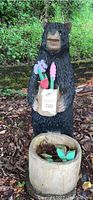 Front view of black and tan wooden bear statue holding wooden flower pot with painted wooden flowers. Wooden planter pot on ground in front.