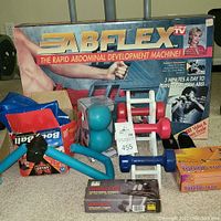 Overall view of exercise equipment lot showing boxed AB Flex abdominal machine, Miracle Balls, Body Ball, hand weights, resistance tubes, and hand grips.