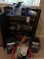 Wooden 3-shelf bookcase filled with various items including a plastic container, glass, two RCA speakers on the floor, shoehorns, and a red basket with papers and small items.