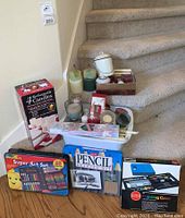 View of the full lot showing candles grouped on stairs and art supplies on floor, including boxed RoseArt Super Art Set, Pencil Drawing Kit, and Painting Case.