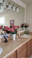Wide bathroom countertop with Lenox vase holding artificial pink and white flowers, additional floral arrangement, clear glass soap dish, framed pictures, glass bottles, sink, and mirror above.