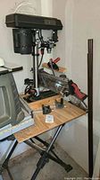 Overall view of the drill press on a wooden stand along with clamps and other miscellaneous items in a storage or workshop area.