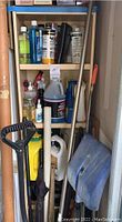 Snow shovel with black plastic scoop leaning against shelving with garden chemicals and tools.