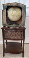 Full view of the 12 inch globe still in its original cardboard box placed on top of the petite wooden side table with a wicker drawer and lower shelf.