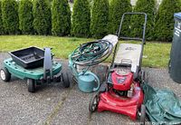 Full lot showing Craftsman red lawn mower with Dust Blocker bag, coiled garden hoses on metal storage container, green watering can, green metal yard cart with black wheels, and the folded tarp