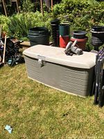 Large beige Rubbermaid storage box with lid placed on grass surrounded by planting pots and other containers. Circular saw is on top of the storage box.