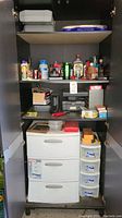 Photo of black storage shelf interior showing three shelves and three plastic drawers with assorted tools, bottles, and boxes.