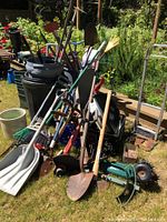 Photo of various garden tools including shovels, spades, rakes, broom, and garbage cans stacked outdoors on grass.