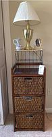Wicker storage bin cabinet with three baskets, lamp, vase, and clock arranged on top.