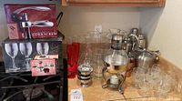 Photo of silver-toned glass coffee pot with stand and assorted glassware including shakers, mugs, shots, and coasters on a kitchen counter