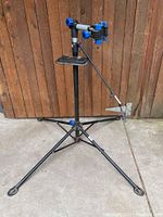 Full view of bike repair stand showing tripod base, upright pole, clamp with blue adjustment knobs, and attached tool tray.
