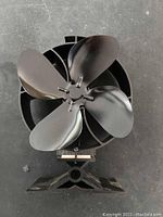 Black heat powered stove fan with four blades, showing the front view of the fan and base.