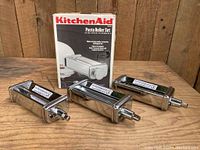 Three KitchenAid pasta attachments including sheet roller, fettuccine and linguine cutters with original box in background