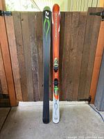 Shows front view of the pair of skis standing upright, one ski topsheet showing orange and white with graphics, other ski base in black with green Rossignol logo.
