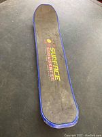 Top view of the Surface snow skate showing the black textured surface with Surface logo in yellow and red and blue edges.
