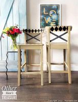 Full view of the two beige barstools with black diamond pattern on backrests, positioned side by side on wood floor beneath framed artwork.