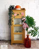 Full view of mustard yellow wooden cabinet with tropical flamingo and leaf decorations, glass front door, staged with plants and flamingo figurine.