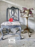 Photo showing refinished dining chair painted in grey and black shades with floral patterned fabric seat. Chair is styled in antique design with carved backrest and cabriole legs. Positioned next to a wooden stand table for scale and staging.