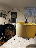 Image showing a large arc floor lamp with a yellow drum lampshade positioned near a bed in a room with framed pictures on the wall and sheer curtains on the window.