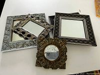 View of four mirrors stacked with the front featuring a small round mirror with ornamental metal frame and tarnished glass, and three larger rectangular/square mirrors with silver and black patterned frames.