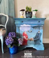 Front view of large blue wooden night stand with hand-painted design showing a couple holding hands under clouds. Three drawers visible, two with brass handles and one with blue knobs. Blue and purple flowers placed next to the stand.