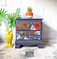 Wooden nightstand painted in purple and gray with large colorful floral designs on front panels and drawers. Includes three drawers with decorative knobs and scalloped base. Nightstand measures 24x23x14 inches.