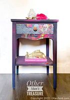 Photo of a refinished wooden night table with painted pink and black surfaces, featuring a drawer decorated with colorful decoupage paper showing floral and skull designs. Lower shelf visible. Table stands on dark wood floor.